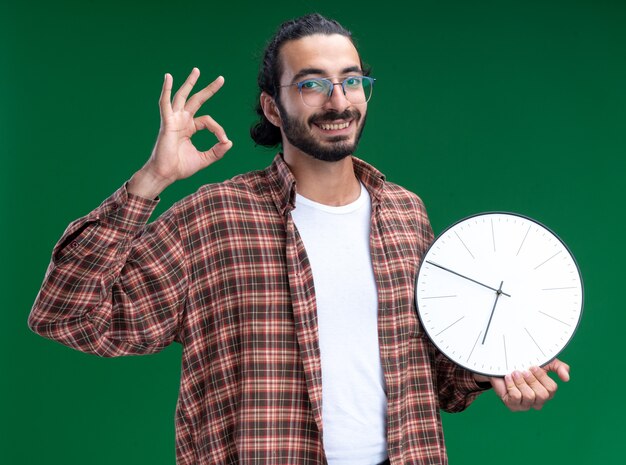 Jovem, bonito, cara de limpeza sorridente, vestindo uma camiseta segurando um relógio de parede, mostrando um gesto de ok isolado na parede verde