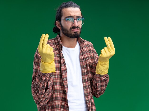 Jovem, bonito, cara de limpeza triste usando camiseta e luvas, mostrando gesto de ponta isolado na parede verde