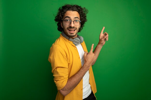 Jovem, bonito, caucasiano, impressionado, usando óculos com fones de ouvido ao redor do pescoço, em vista de perfil, olhando para a câmera apontando para cima, isolado em um fundo verde com espaço de cópia