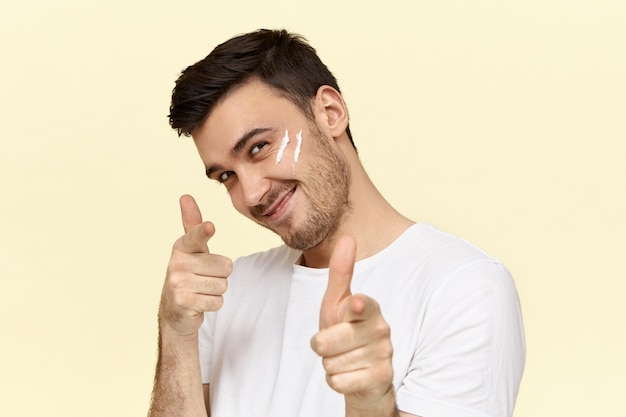 Jovem bonito com a barba por fazer em uma camiseta branca sorrindo e apontando o dedo indicador para a frente