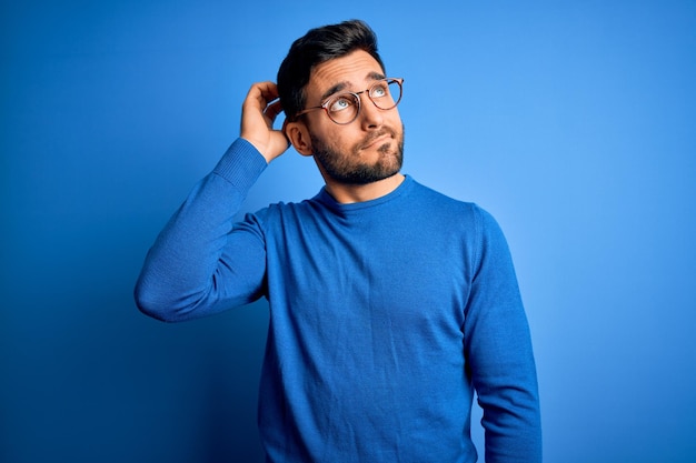 Jovem bonito com barba, vestindo suéter casual e óculos sobre fundo azul, confunde e se pergunta sobre a questão incerta com dúvida pensando com a mão na cabeça conceito pensativo