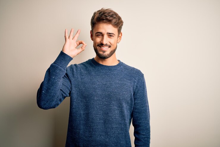 Foto grátis jovem bonito com barba vestindo suéter casual em pé sobre fundo branco sorrindo positivo fazendo sinal de ok com a mão e os dedos expressão de sucesso