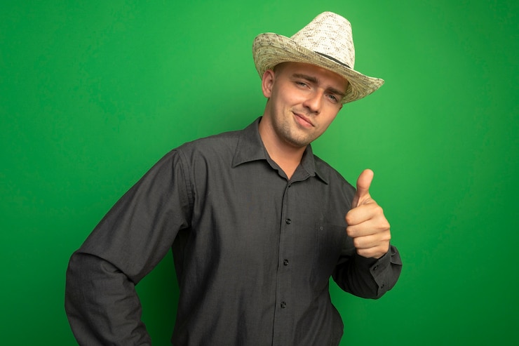 Foto grátis jovem bonito com camisa cinza e chapéu de verão, olhando para a frente, sorrindo com uma cara feliz e mostrando os polegares em pé sobre a parede verde