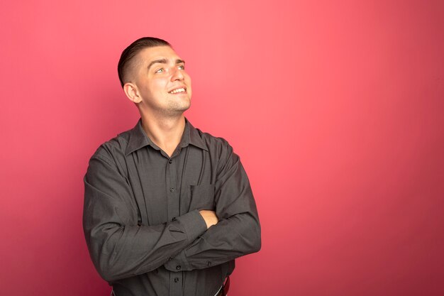 Jovem bonito com camisa cinza olhando para o lado com um sorriso no rosto e os braços cruzados sobre o peito em pé sobre a parede rosa