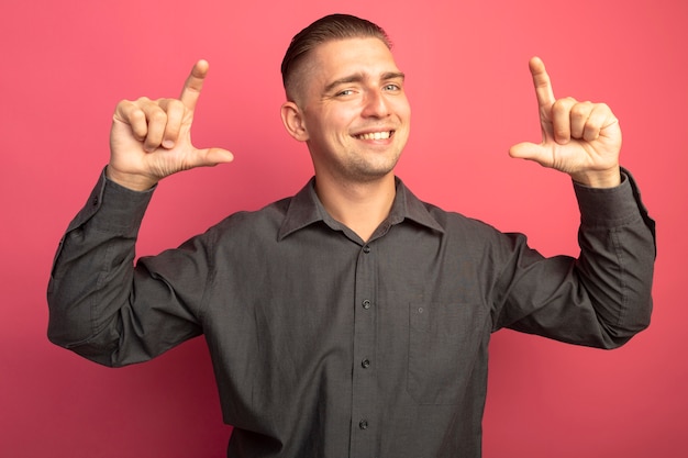 Foto grátis jovem bonito com camisa cinza sorrindo alegremente mostrando o gesto do tamanho com os dedos medindo o símbolo em pé sobre a parede rosa