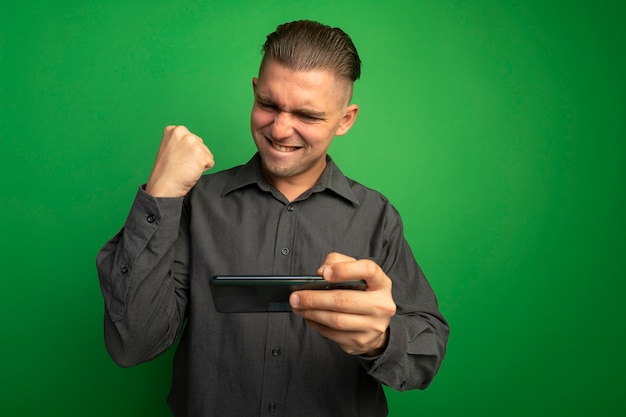 Jovem bonito com camisa cinza usando o gadget, punho cerrado, feliz e animado, regozijando-se com seu sucesso em pé sobre a parede verde