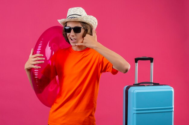 Jovem bonito com camiseta laranja e chapéu de verão, usando óculos escuros pretos segurando um anel inflável, fazendo um gesto de me ligar com a mão positiva e feliz em pé com a mala de viagem sobre rosa