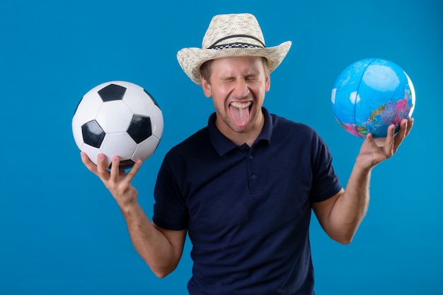 Jovem bonito com chapéu de verão segurando uma bola de futebol e um globo louco feliz gritando fascinado, mostrando a língua em pé sobre um fundo azul