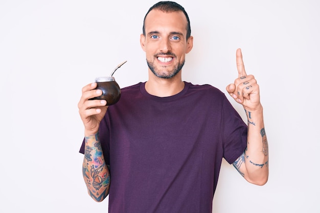 Jovem bonito com tatuagem bebendo infusão de mate sorrindo com uma ideia ou pergunta apontando o dedo com cara feliz, número um