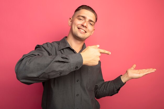 Jovem bonito com uma camisa cinza apresentando algo com o braço das mãos apontando com o dedo indicador para o lado, sorrindo confiante em pé sobre a parede rosa