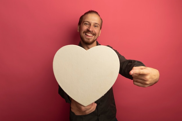 Foto grátis jovem bonito com uma camisa cinza segurando um coração de papelão apontando com o dedo indicador para ele e sorrindo com uma cara feliz