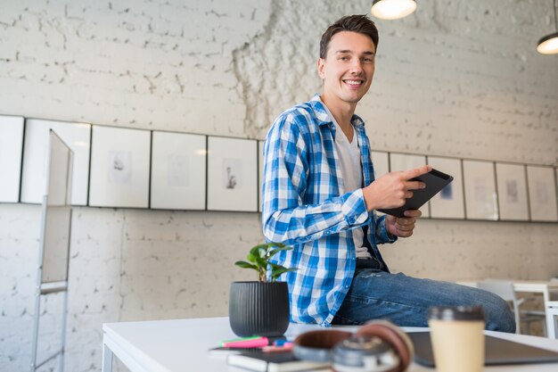 Jovem bonito com uma camisa de bochecha sentado na mesa usando um computador tablet em um escritório colaborativo