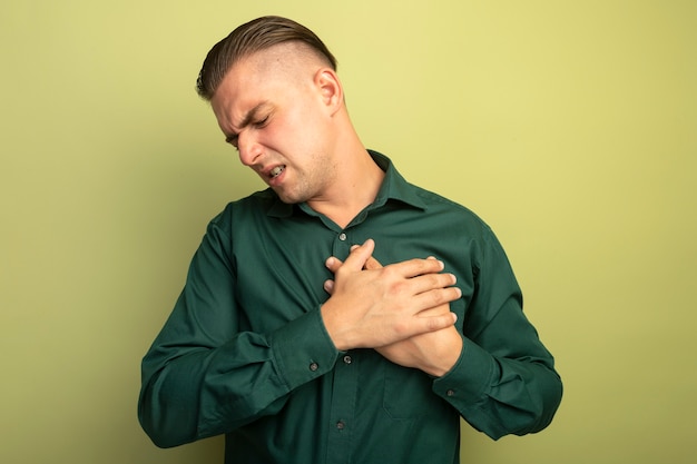 Foto grátis jovem bonito com uma camisa verde parecendo doente, segurando a mão no peito, sentindo dor em pé sobre a parede de luz