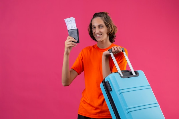 Jovem bonito com uma camiseta laranja em pé com uma mala de viagem segurando as passagens aéreas, sorrindo alegremente sobre o fundo rosa