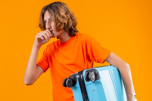 Jovem bonito com uma camiseta laranja segurando uma mala de viagem olhando de lado e sorrindo confiante em pé sobre um fundo amarelo