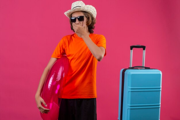 Jovem bonito com uma camiseta laranja usando óculos escuros pretos segurando um anel inflável em pé com uma mala de viagem parecendo surpreso cobrindo a boca com a mão sobre o fundo rosa