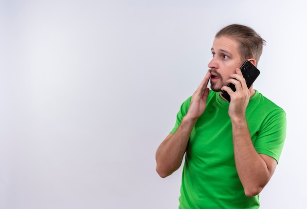 Jovem bonito com uma camiseta verde falando no celular parecendo surpreso e surpreso em pé sobre um fundo branco