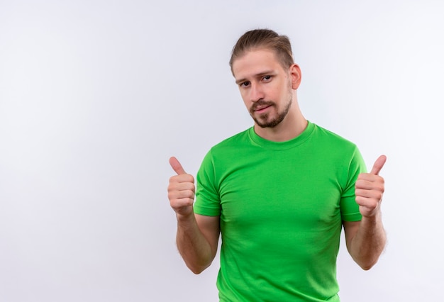 Jovem bonito com uma camiseta verde olhando para a câmera e sorrindo amigável mostrando os polegares em pé sobre um fundo branco