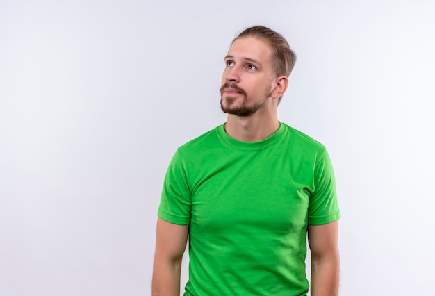 Jovem bonito com uma camiseta verde olhando para o lado com uma expressão pensativa no rosto em pé sobre um fundo branco