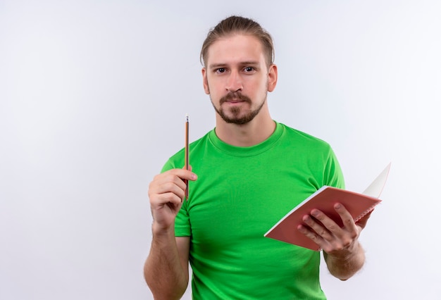 Jovem bonito com uma camiseta verde segurando um caderno apontando com um lápis para cima tendo uma ótima ideia em pé sobre um fundo branco