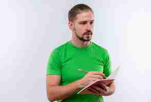 Foto grátis jovem bonito com uma camiseta verde segurando um caderno e escrevendo algo com uma expressão séria no rosto em pé sobre um fundo branco