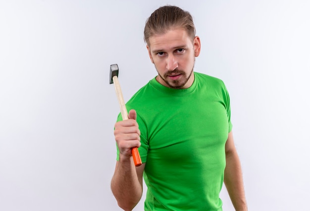 Jovem bonito com uma camiseta verde segurando um martelo, olhando para a câmera com uma cara séria em pé sobre um fundo branco