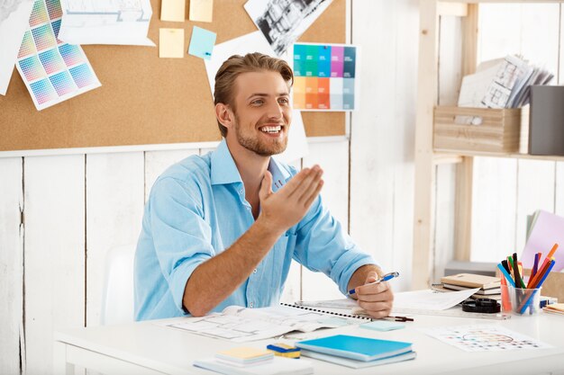 Jovem bonito confiante sorridente empresário trabalhando sentado à mesa. Interior de escritório moderno branco
