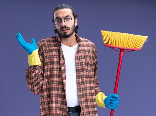 Jovem bonito confuso, cara de limpeza usando camiseta e luvas, segurando o esfregão estendendo a mão isolada na parede azul