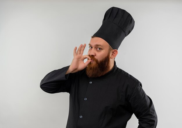 Jovem bonito cozinheiro confiante em uniforme de chef, fazendo gestos saborosos e isolado na parede branca