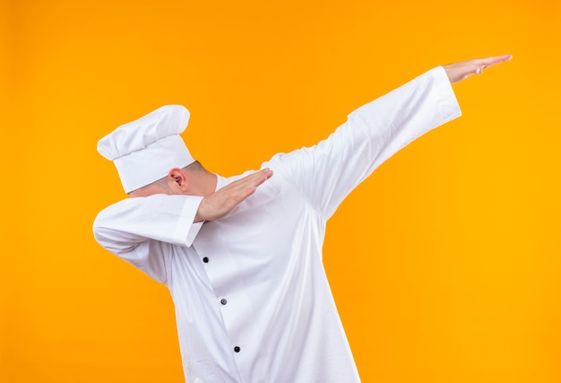 Jovem bonito cozinheiro em uniforme de chef colocando a cabeça no braço e levantando outro braço isolado no espaço laranja