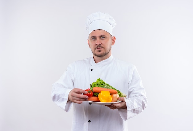 Jovem bonito cozinheiro em uniforme de chef segurando o prato com legumes olhando para o espaço em branco isolado