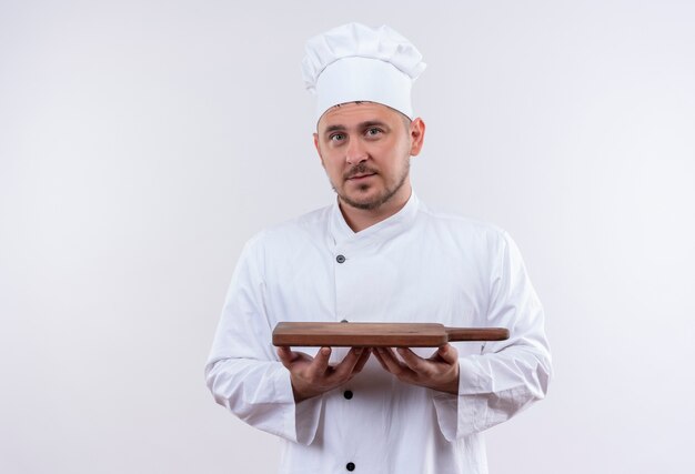 Jovem bonito cozinheiro em uniforme de chef segurando uma tábua de corte e parecendo isolado no espaço em branco