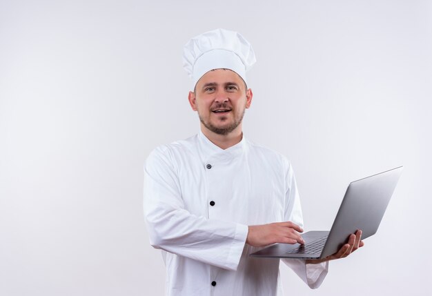 Jovem bonito cozinheiro satisfeito com uniforme de chef segurando laptop em um espaço em branco isolado