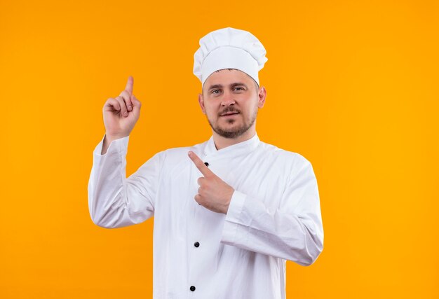 Jovem bonito cozinheiro satisfeito em uniforme de chef apontando para cima isolado no espaço laranja