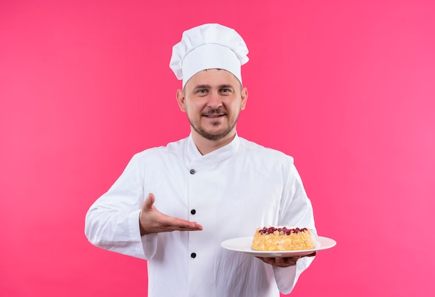 Jovem bonito cozinheiro sorridente em uniforme de chef segurando e apontando para o prato de bolo isolado no espaço rosa