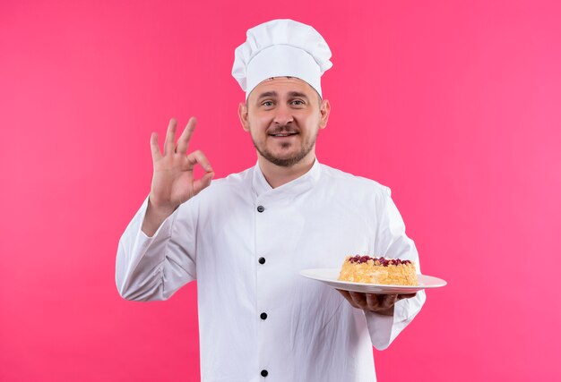 Jovem bonito cozinheiro sorridente em uniforme de chef segurando o prato de bolo fazendo sinal de ok isolado no espaço rosa