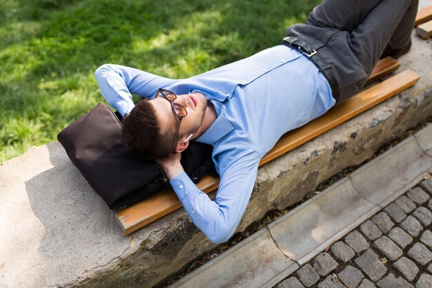 Jovem bonito de camisa azul e óculos de sol com fones de ouvido sem fio sonhadoramente deitado no banco enquanto passa o tempo no parque da cidade