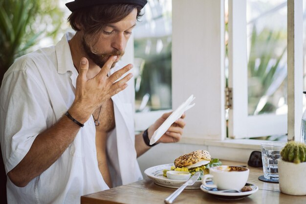 Jovem bonito de camisa branca aberta, tomando café da manhã em um café com um hambúrguer vegetariano, bebendo café, estilo de vida em uma ilha tropical, vida em Bali.