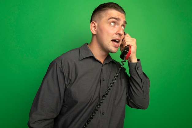 Jovem bonito de camisa cinza falando no telefone vintage, parecendo de lado confuso em pé sobre uma parede verde