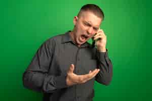 Foto grátis jovem bonito de camisa cinza gritando com expressão agressiva enquanto fala no celular, em pé sobre a parede verde