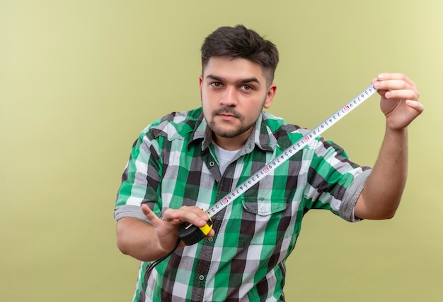 Jovem bonito de camisa quadriculada segurando um medidor em pé sobre uma parede cáqui