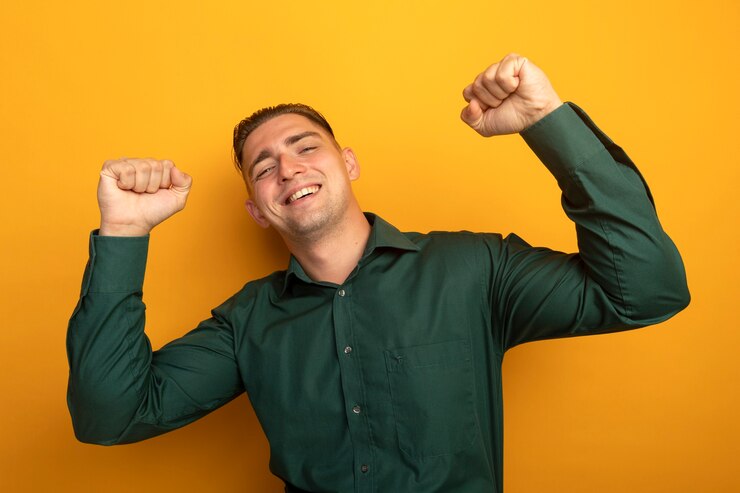Foto grátis jovem bonito de camisa verde cerrando os punhos, feliz e animado, regozijando-se com o sucesso em pé sobre a parede laranja