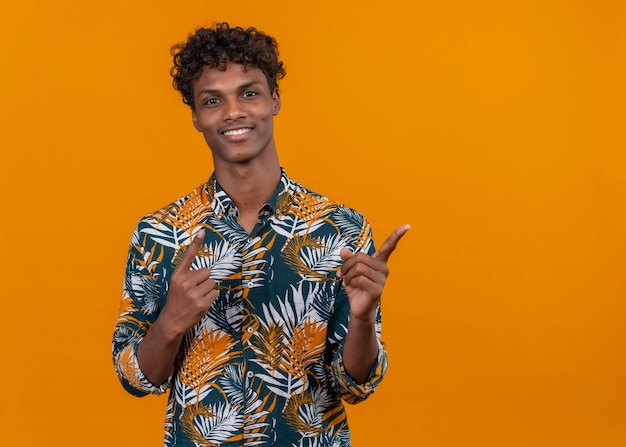 Jovem bonito de pele escura sorridente com cabelo encaracolado em uma camisa estampada de folhas apontando com o dedo indicador para cima enquanto olha para a câmera em um fundo laranja