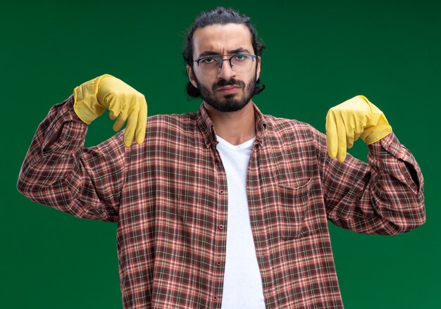 Jovem, bonito, descontente, faxineiro, usando camiseta e luvas, fingindo que está segurando algo isolado na parede verde