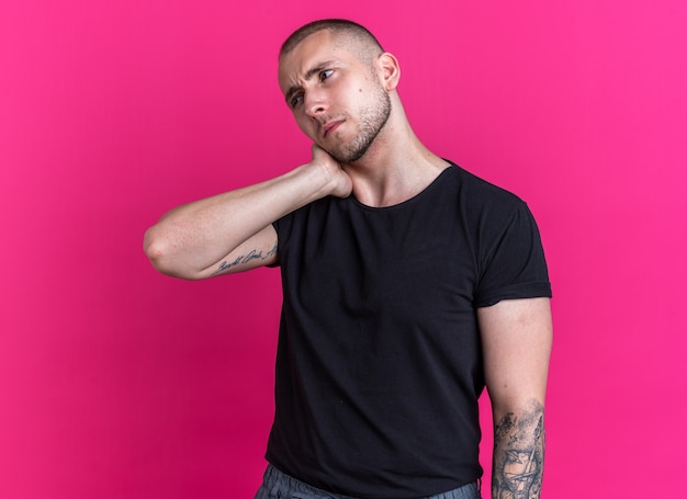 Foto grátis jovem bonito descontente vestindo uma camiseta preta e colocando a mão no pescoço dolorido isolado na parede rosa