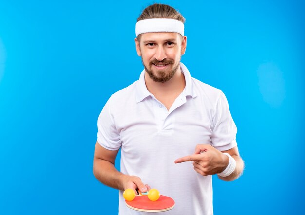 Jovem, bonito, desportivo, sorridente, usando bandana e pulseiras, segurando e apontando para uma raquete de pingue-pongue com bolas isoladas no espaço azul