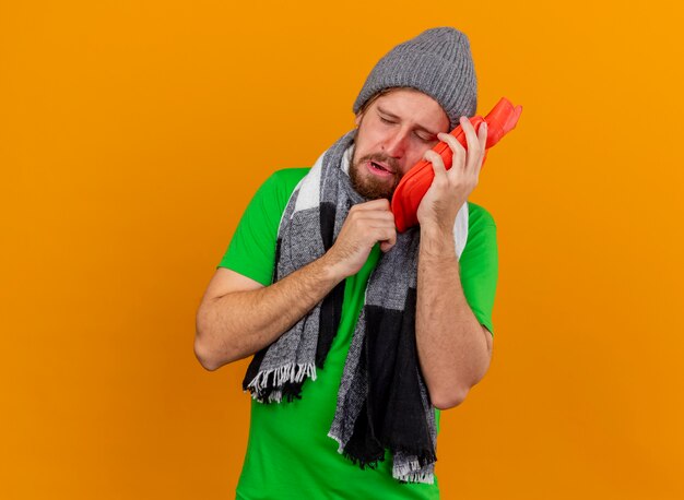 Jovem bonito doente, dolorido, usando chapéu de inverno e lenço segurando uma bolsa de água quente tocando o rosto com os olhos fechados, isolado na parede laranja