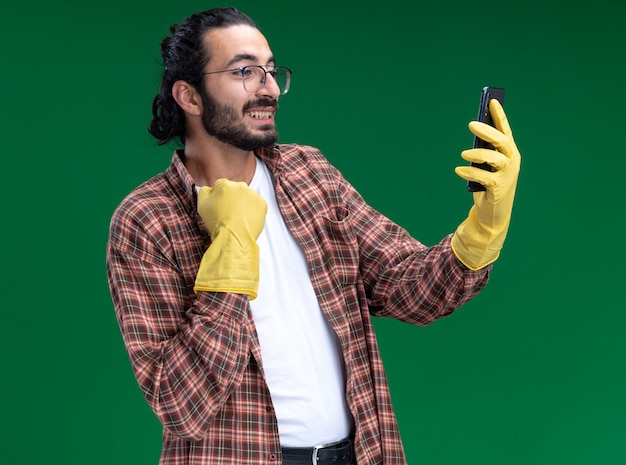 Jovem, bonito e aquecido, cara de limpeza usando camiseta e luvas, tira uma selfie agarrada na gola isolada na parede verde