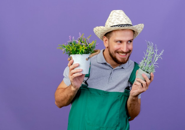 Jovem bonito e astuto jardineiro eslavo de uniforme e chapéu segurando vasos de flores, olhando para o lado e sorrindo, isolado na parede roxa com espaço de cópia