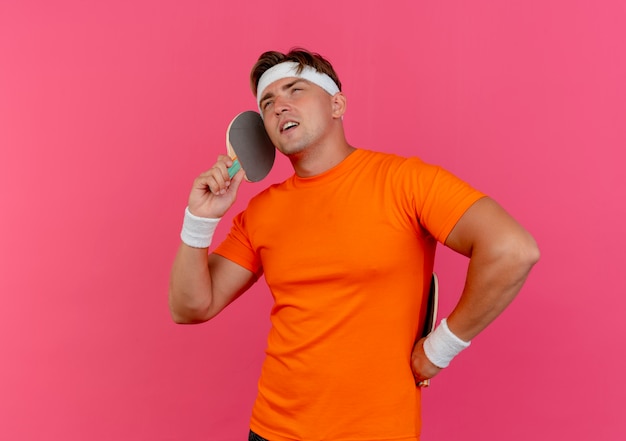 Jovem bonito e confiante homem desportivo usando bandana e pulseiras segurando raquetes de pingue-pongue e fingindo estar falando no telefone isolado no rosa com espaço de cópia